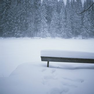Valokuva lumeen hautautuneesta penkistä järven rannalla. Riitta Supperin valokuvavedos.